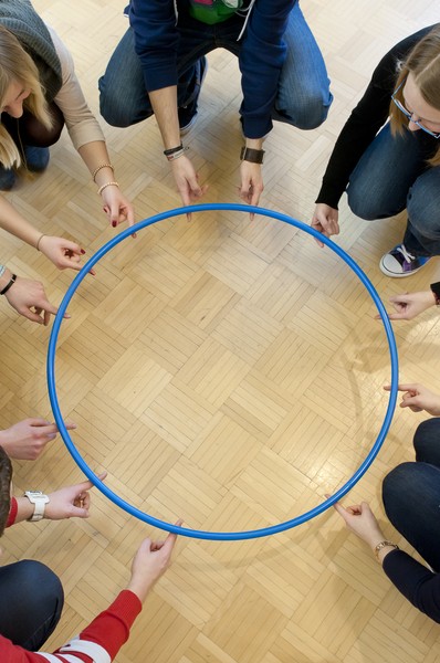 Sechs Kursteilnehmende heben gemeinsam einen Hula Hoop-Ring mit den Zeigefingern vom Boden in die Höhe.