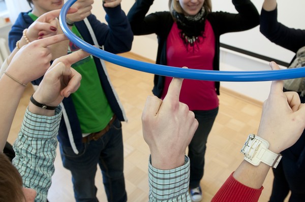 Eine Gruppe Menschen hält gemeinsam mit den Zeigefingern eine Hula Hoop Ring auf.